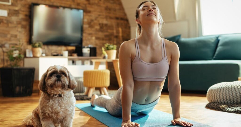 Young athletic woman in cobra pose practicing Yoga with her dog at home.