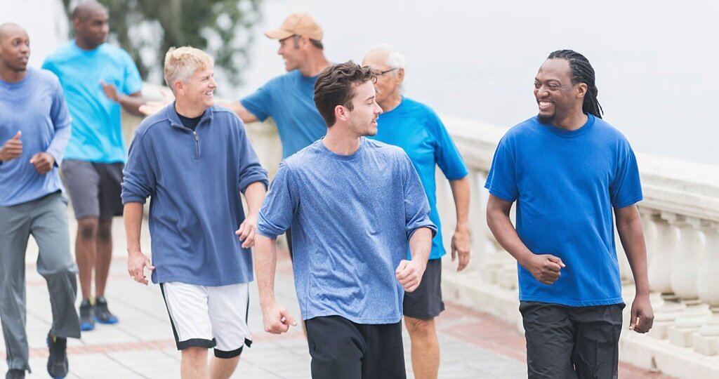 Multiracial group of men wearing blue shirts, jogging or powerwalking in the park along the waterfront. Focus is on the two men talking in the foreground.