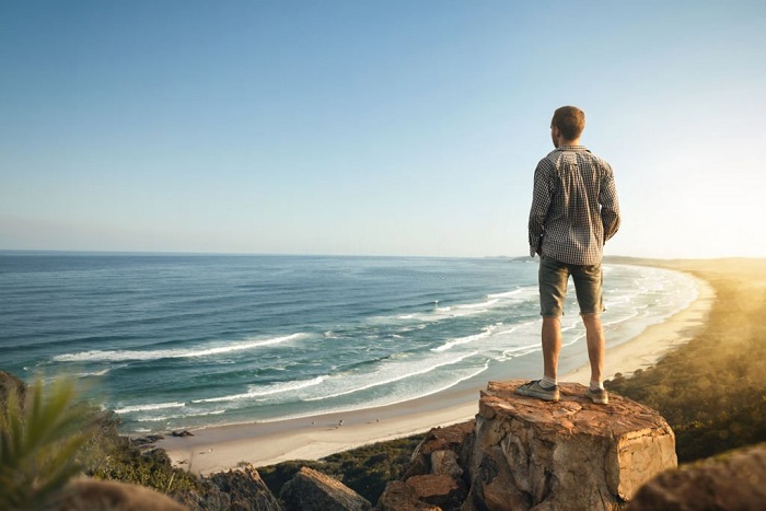 Rear view of Avery standing on top of a rock overlooking a vast beach while the sun is low