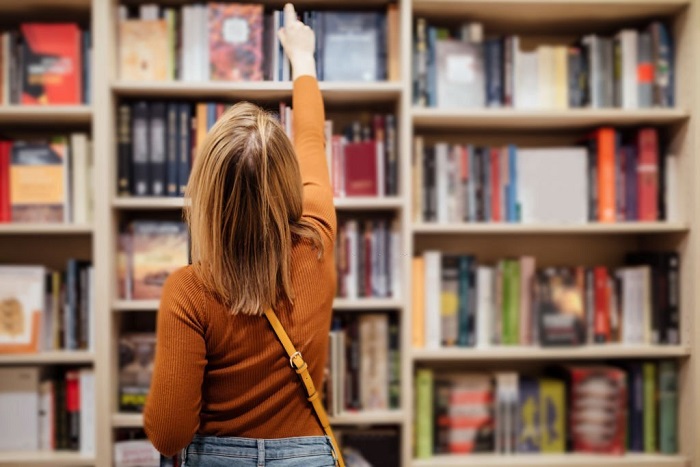 Eden with loose long blondie hair standing and holding an open book between book shelves in the library