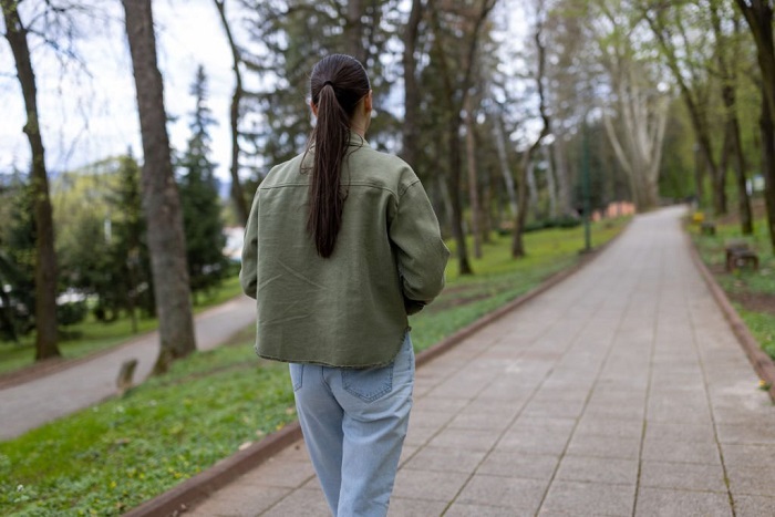 Rear view of Haley slowly walking through alley of green trees on sidewalk at city park