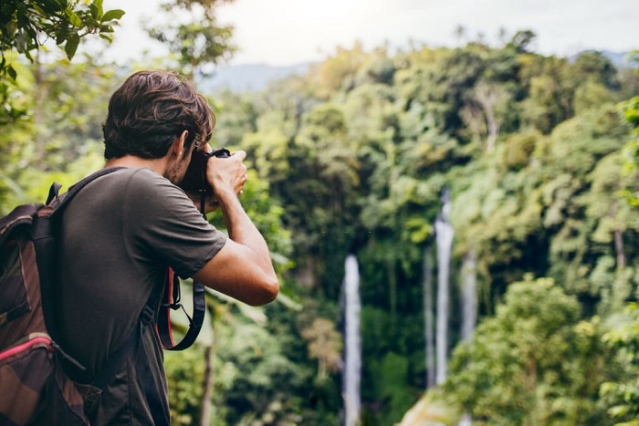 Man with backpack standing in front of waterfall and taking a photo