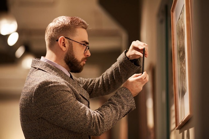 Side view of Parker in eyeglasses taking photo of paintings on his mobile phone in the gallery